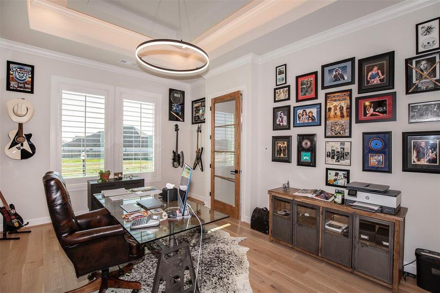 This stylish home office features a glass desk, leather chair, and ample natural light from large windows with plantation shutters. The walls are adorned with a collection of framed music memorabilia, and the room is accented with guitars, creating a modern and creative workspace.