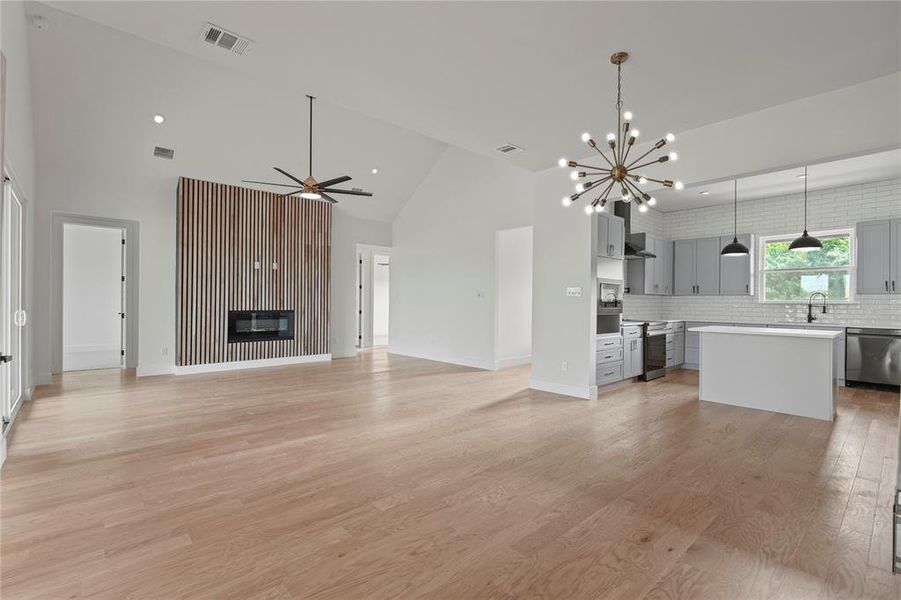 Unfurnished living room with high vaulted ceiling, ceiling fan, a chandelier, light wood finished floors, and a glass covered fireplace Unfurnished living room with high vaulted ceiling, ceiling fan, a chandelier, light wood finished floors, and a glass covered fireplace