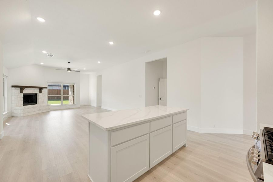 Open-concept kitchen with white island and quartz countertop overlooking living room with stone fireplace and light wood floors Open-concept kitchen with white island and quartz countertop overlooking living room with stone fireplace and light wood floors