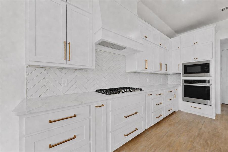 Contemporary kitchen featuring white cabinetry with gold-tone hardware, a white herringbone tile backsplash, and light-colored countertops Contemporary kitchen featuring white cabinetry with gold-tone hardware, a white herringbone tile backsplash, and light-colored countertops