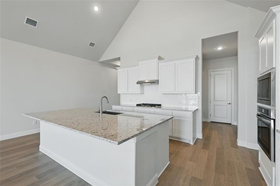 Kitchen with light stone counters, a kitchen island with sink, stainless steel appliances, vaulted ceiling, and white cabinets