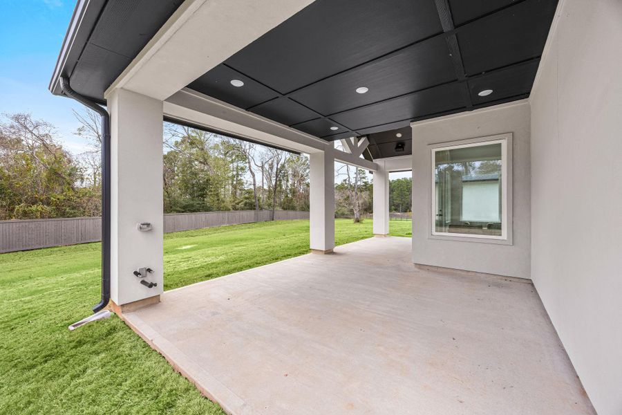 A spacious covered patio with a modern design, featuring a black ceiling and recessed lighting. It overlooks a large, well-maintained backyard perfect for outdoor relaxation and entertainment.