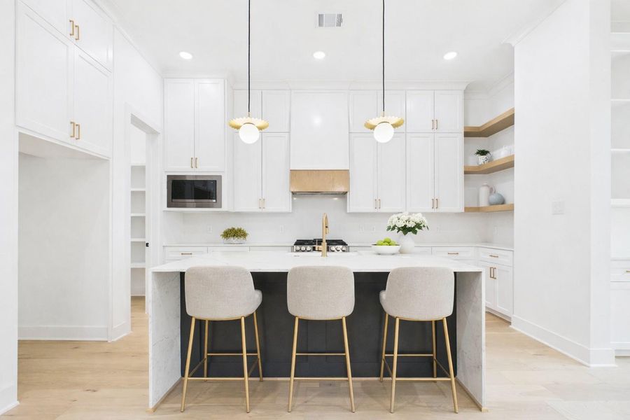 Bright and spacious kitchen with modern design, featuring sleek white cabinetry, a large central island with a dark accent, and elegant pendant lighting. The space includes stainless steel appliances, ample natural light from large windows, and light wood flooring, creating a welcoming and functional environment.