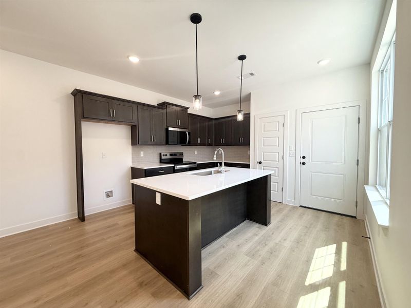 Kitchen featuring dark wood finish cabinetry, stainless steel appliances, light wood-style floors, an island with sink, and decorative light fixtures