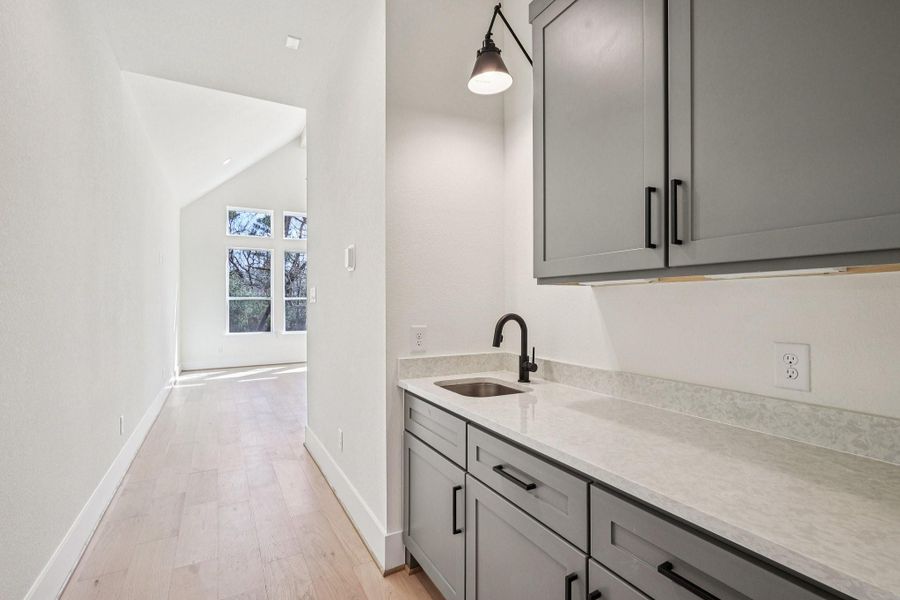 This photo features a bright hallway with light wood flooring leading to large windows, offering plenty of natural light. On the right, there's a sleek wet bar with gray cabinets, a modern black faucet, and a single pendant light.