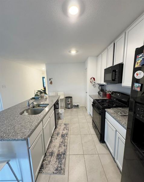 Kitchen with white cabinets, black appliances, an island with sink, and light stone counters