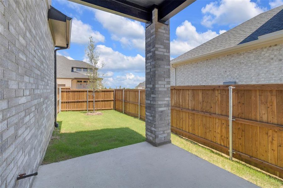 Covered concrete patio with brick column, overlooking a fenced backyard with a small tree and green lawn