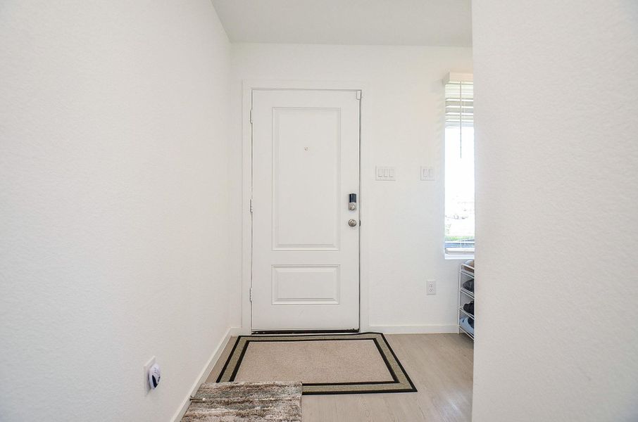 This photo features a modern kitchen with white cabinetry, stainless steel appliances, and a spacious island. The open layout flows into a bright entryway with light wood flooring, creating a welcoming ambiance.