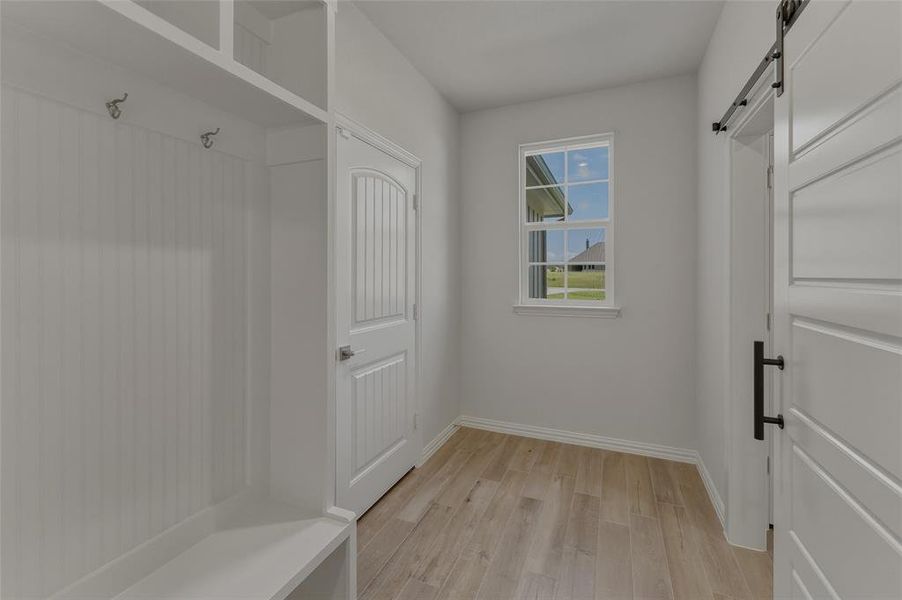 Built-in bench with hooks and upper shelving, plank wood-finish flooring, white trim, sliding barn door with black hardware, and double-hung window