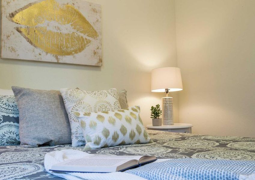 Staged bedroom with painting that has golden lips, bed with multiple pillows and blue comforter and light in the corner.