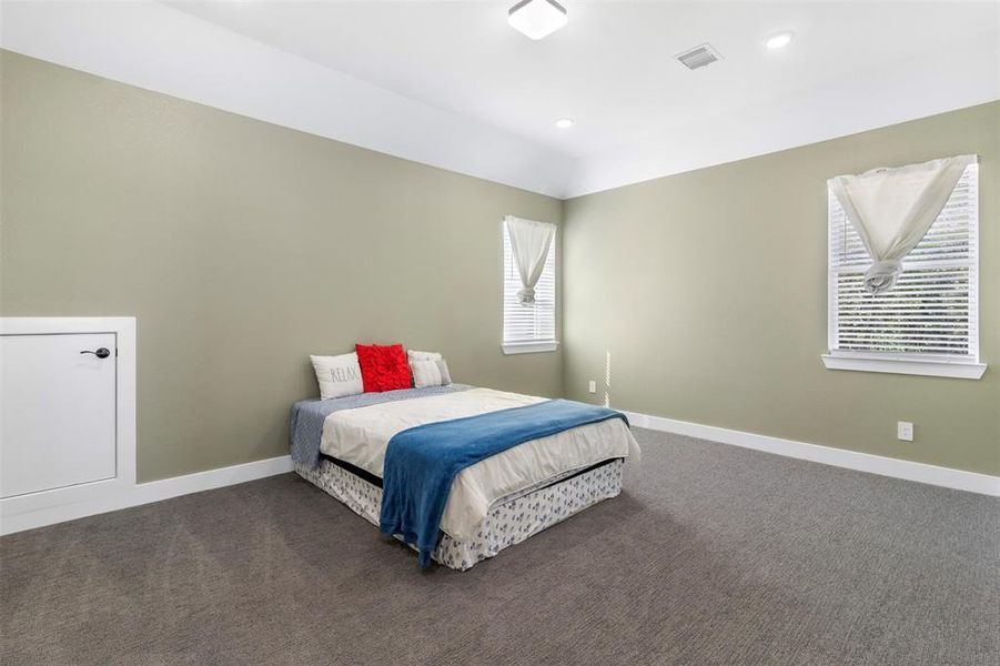 This spacious bedroom features soft green walls, carpeted flooring, and ample natural light from two windows. It includes a cozy bed with simple decor, creating a relaxing atmosphere. Ideal for comfort and tranquility.