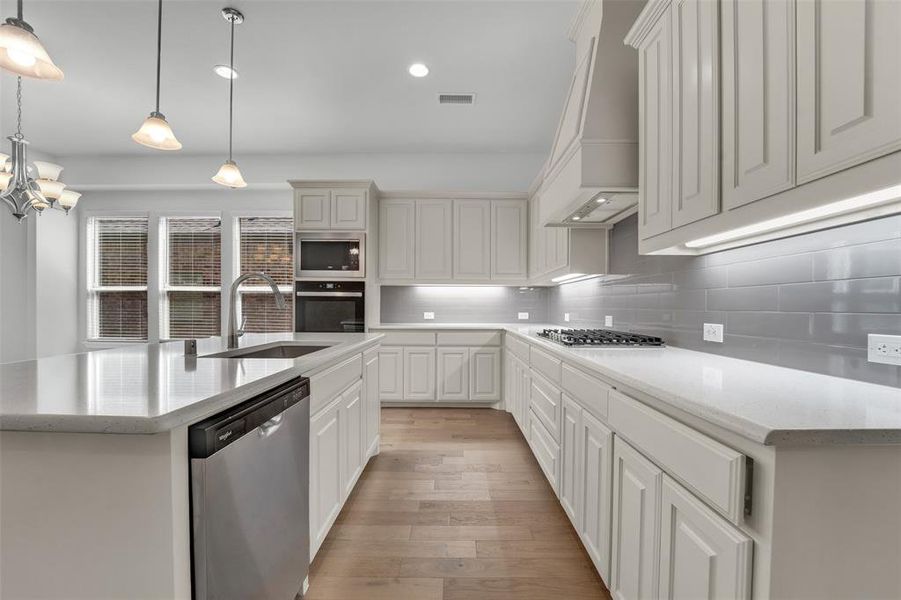 Kitchen with stainless steel appliances, light stone counters, a center island with sink, light wood finished floors, and tasteful backsplash