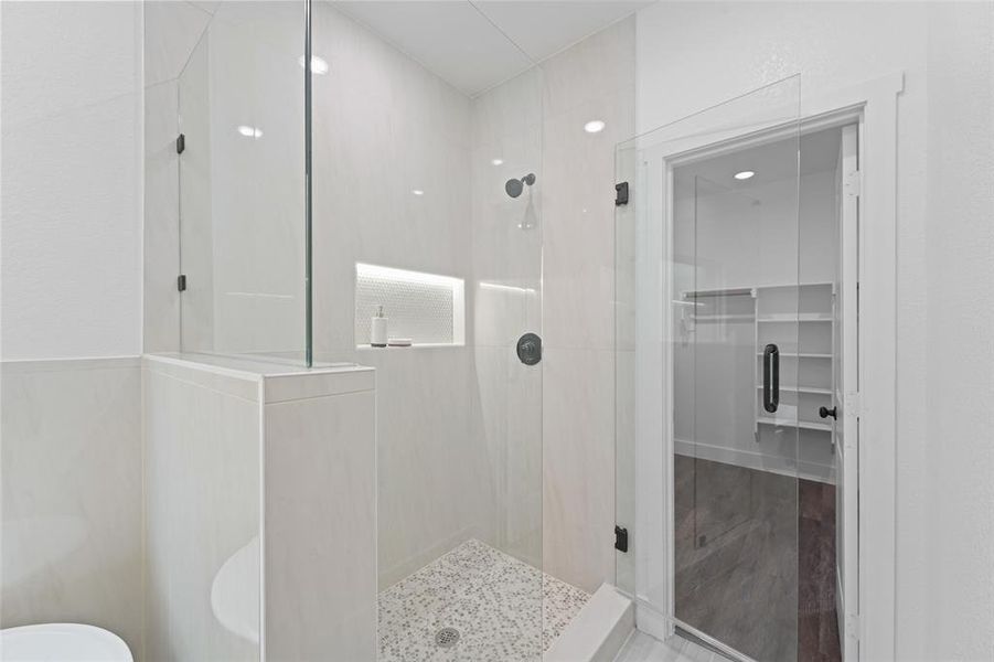 Walk-in shower featuring a frameless glass enclosure, tiled walls, built-in shelving with accent lighting, dark-finish fixtures, and a pebble-mosaic floor Walk-in shower featuring a frameless glass enclosure, tiled walls, built-in shelving with accent lighting, dark-finish fixtures, and a pebble-mosaic floor
