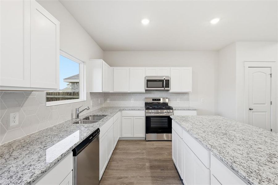 Kitchen featuring white shaker cabinetry, stainless steel appliances, light-toned granite countertops, a hexagonal tile backsplash, and wood-finish flooring Kitchen featuring white shaker cabinetry, stainless steel appliances, light-toned granite countertops, a hexagonal tile backsplash, and wood-finish flooring