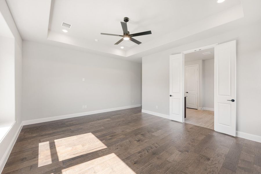 Empty bedroom with tray ceiling, ceiling fan, dark hardwood floors, and open double doors Empty bedroom with tray ceiling, ceiling fan, dark hardwood floors, and open double doors