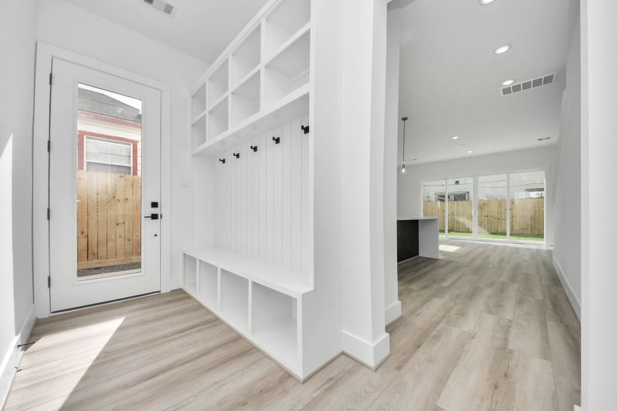Bright entryway with built-in storage and hooks, leading to an open-concept living area with large windows overlooking a fenced backyard. Light wood flooring and modern finishes throughout.