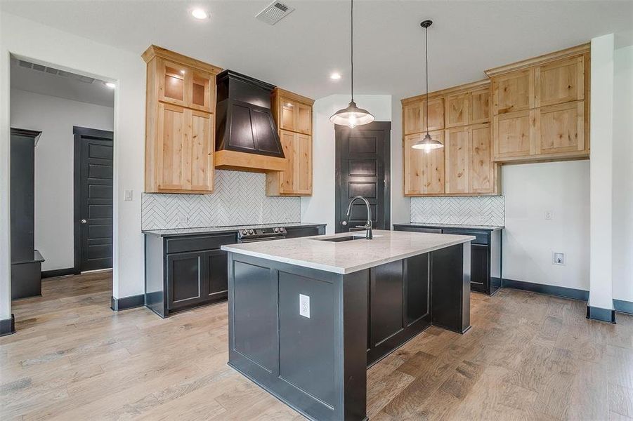 Kitchen featuring backsplash, custom exhaust hood, an island with sink, light wood-type flooring, and sink