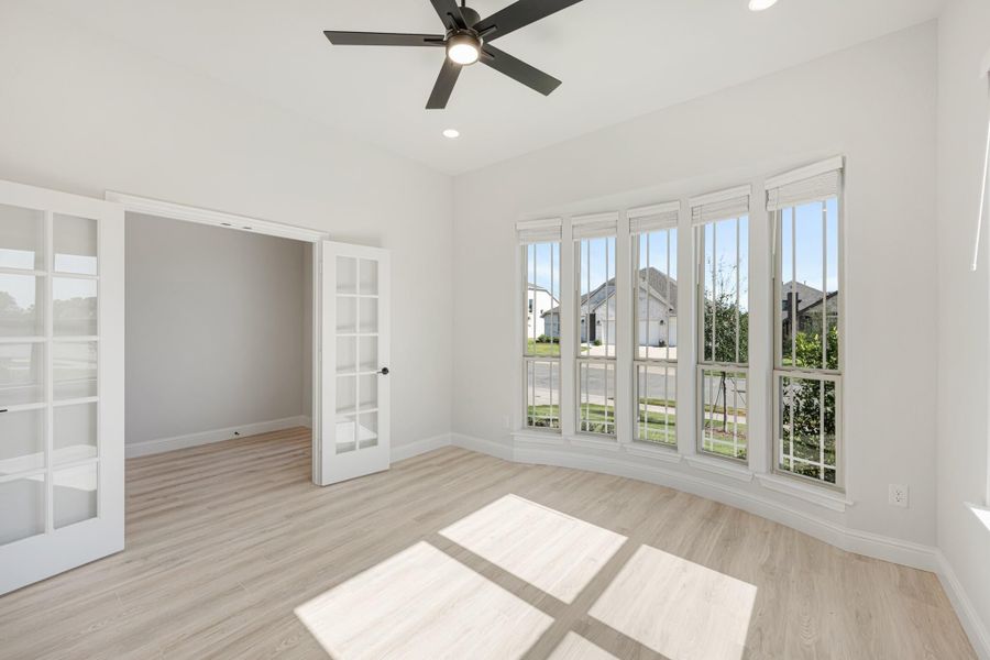 Empty room with light wood floors, ceiling fan, large windows, and French doors leading to adjacent space
