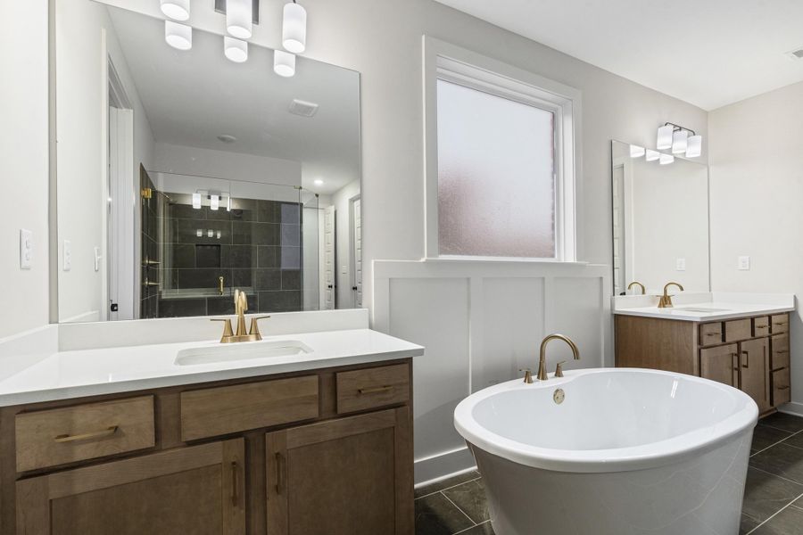 Freestanding soaking tub with brushed gold fixtures, dual wood-finish vanities with white countertops, and a tiled shower enclosure visible in the mirror's reflection Freestanding soaking tub with brushed gold fixtures, dual wood-finish vanities with white countertops, and a tiled shower enclosure visible in the mirror's reflection