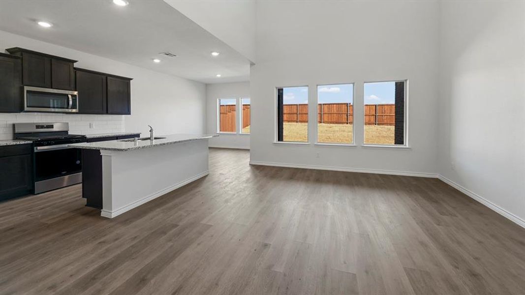 Open-concept living area featuring wood-finish flooring, a kitchen island with a sink, dark cabinetry, stainless steel appliances, and a white subway tile backsplash Open-concept living area featuring wood-finish flooring, a kitchen island with a sink, dark cabinetry, stainless steel appliances, and a white subway tile backsplash
