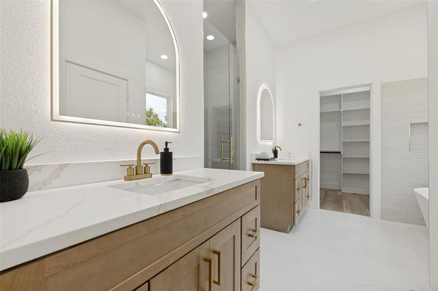 Spacious bathroom featuring a double vanity with stone countertops, modern gold-finish faucets, illuminated arched mirrors, a glass-enclosed shower, and a large walk-in closet with built-in shelving Spacious bathroom featuring a double vanity with stone countertops, modern gold-finish faucets, illuminated arched mirrors, a glass-enclosed shower, and a large walk-in closet with built-in shelving
