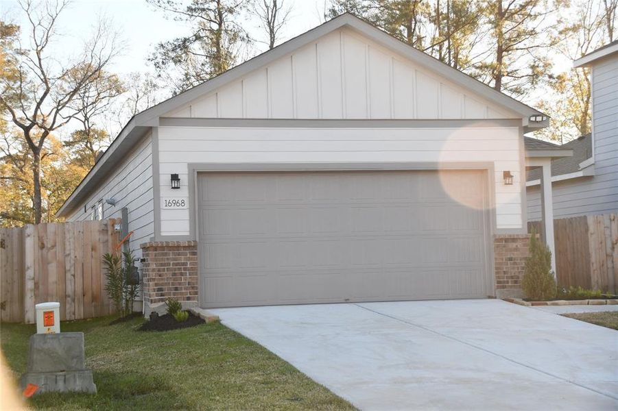 This photo shows a modern single-story home with a two-car garage. It features a clean exterior with a combination of light-colored siding and brick accents. The driveway is spacious, and the property is bordered by a wooden fence. The yard is modestly landscaped with a few shrubs and trees in the background, offering a tranquil setting.