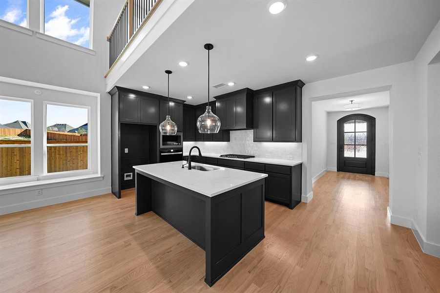 Kitchen featuring dark cabinetry, decorative light fixtures, an island with sink, light wood-type flooring, and decorative backsplash Kitchen featuring dark cabinetry, decorative light fixtures, an island with sink, light wood-type flooring, and decorative backsplash