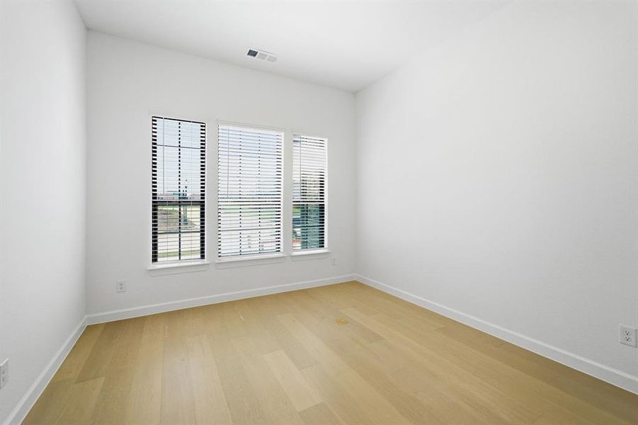 Bright interior room featuring three windows with blinds, wood-finish flooring, and white baseboards Bright interior room featuring three windows with blinds, wood-finish flooring, and white baseboards