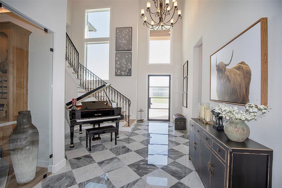 This elegant foyer features a high ceiling with a grand chandelier, large windows for natural light, and a sophisticated black and white tile floor. A sleek piano adds a touch of luxury, complemented by modern artwork and a stylish console with decorative accents.