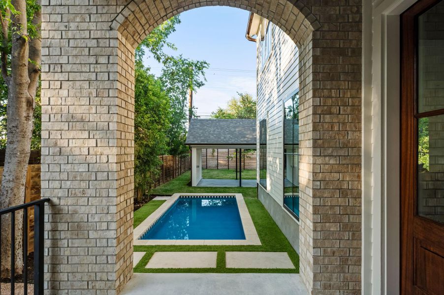 An inviting brick archway reveals a private backyard oasis with a refreshing pool, ample green space, and a covered patio structure.