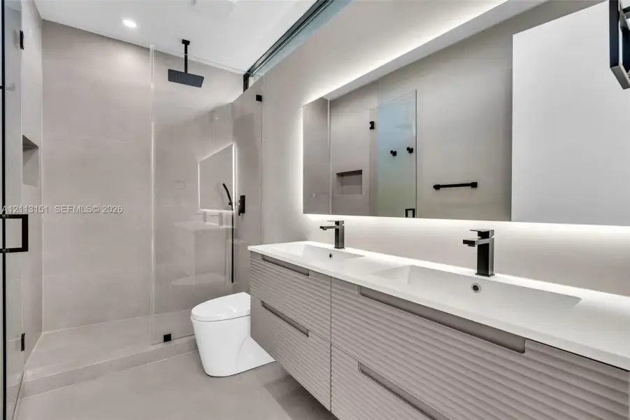 Floating double vanity with textured cabinetry, integrated sinks, and matte black fixtures, paired with a frameless glass shower enclosure featuring an overhead rainfall showerhead and wall-mounted hand shower, finished with large-format stone-tile flooring and an LED-backlit mirror Floating double vanity with textured cabinetry, integrated sinks, and matte black fixtures, paired with a frameless glass shower enclosure featuring an overhead rainfall showerhead and wall-mounted hand shower, finished with large-format stone-tile flooring and an LED-backlit mirror
