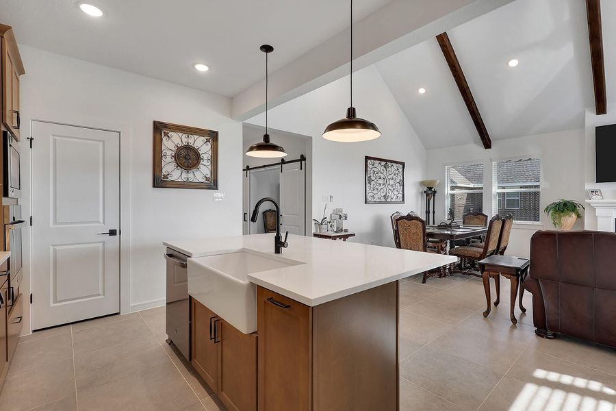 Lovely farmhouse modern style kitchen with open-concept kitchen, living and dining area. It features a large quartz island with a farmhouse sink, dishwasher, pendant lighting, and high ceilings with exposed beams.