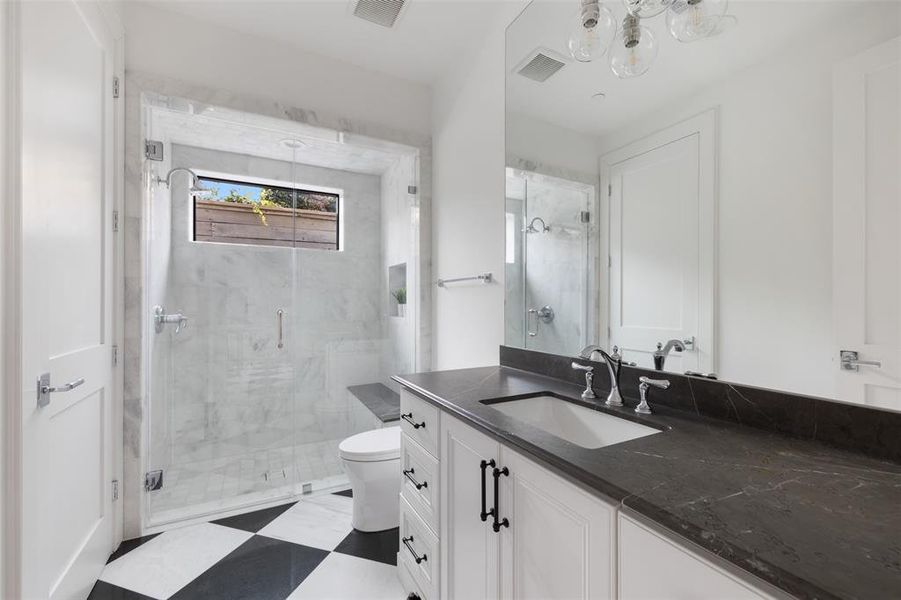 The en suite ground floor bathroom features a stone countertop with furniture-style cabinetry and a frameless glass walk-in shower with bench seating and window. Image from previously completed home in the community.