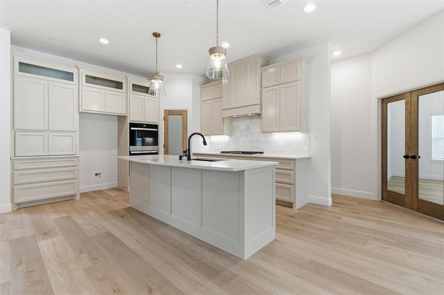 Kitchen featuring decorative backsplash, hanging light fixtures, light wood finished floors, a center island with sink, and light stone countertops Kitchen featuring decorative backsplash, hanging light fixtures, light wood finished floors, a center island with sink, and light stone countertops