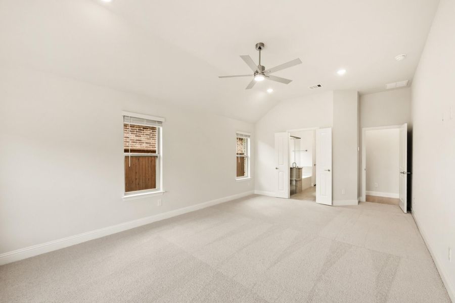 Spacious empty bedroom with vaulted ceiling, ceiling fan, carpet flooring, and open doors to ensuite bathroom and closet. Spacious empty bedroom with vaulted ceiling, ceiling fan, carpet flooring, and open doors to ensuite bathroom and closet.
