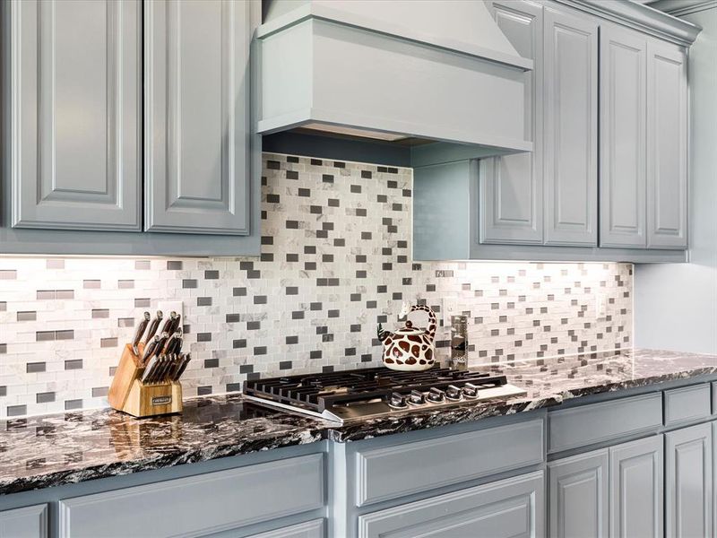 Kitchen with dark stone countertops, premium range hood, tasteful backsplash, and stainless steel gas cooktop Kitchen with dark stone countertops, premium range hood, tasteful backsplash, and stainless steel gas cooktop