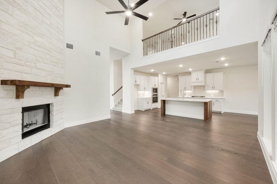 Open living area with stone fireplace, dark hardwood floors, high ceilings, and open kitchen with white cabinets visible in background