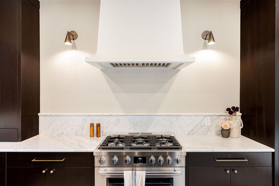 Kitchen with range hood, dark brown cabinets, stainless steel range with gas cooktop, and light stone counters