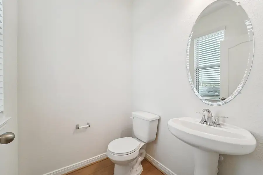 This main-level half bath features a pedestal vanity, oval mirror, window with privacy blinds, modern fixtures, and vinyl plank flooring.