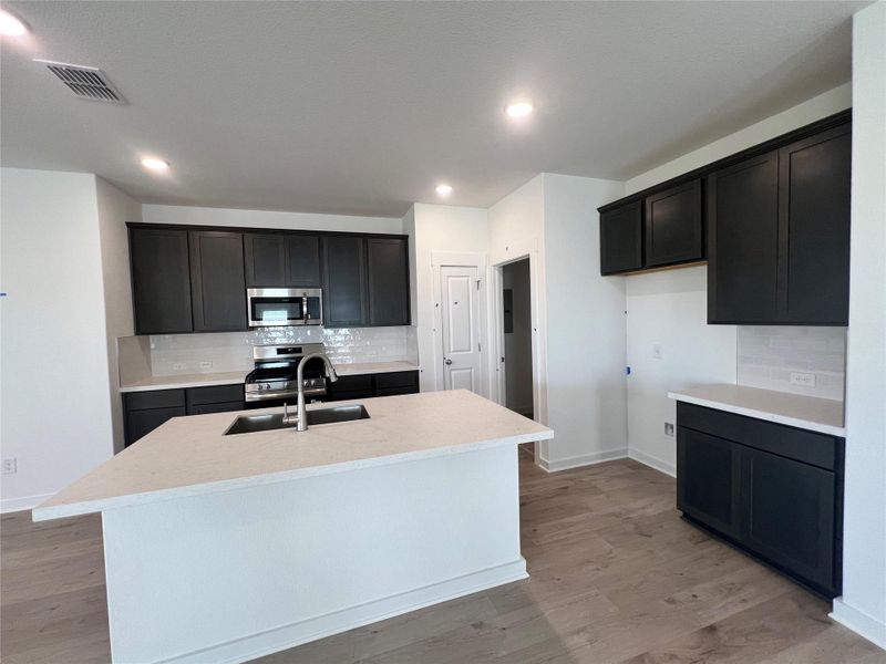 Kitchen with tasteful backsplash, a center island with sink, stainless steel appliances, light wood finished floors, and dark cabinets Kitchen with tasteful backsplash, a center island with sink, stainless steel appliances, light wood finished floors, and dark cabinets