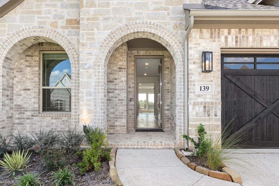 Home entrance with arched brick surround, open front door, and dark wood garage door with landscaped walkway