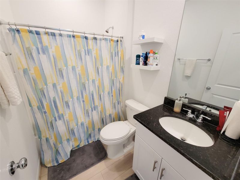 This bathroom features a modern design with a black granite countertop, white vanity, and a colorful shower curtain. It includes a standard toilet, shelves for storage, and a large mirror. The space is well-lit and has a clean, fresh appearance.
