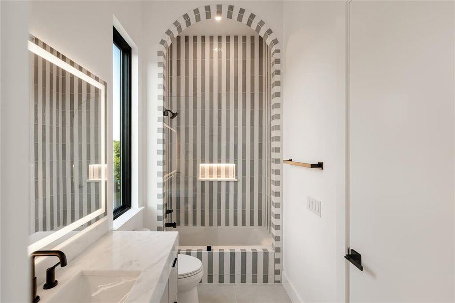 Bathroom featuring a striped tile tub/shower surround with an arched entry, a built-in illuminated niche, a contemporary vanity with an integrated sink, a rectangular backlit mirror, and matte black fixtures Bathroom featuring a striped tile tub/shower surround with an arched entry, a built-in illuminated niche, a contemporary vanity with an integrated sink, a rectangular backlit mirror, and matte black fixtures