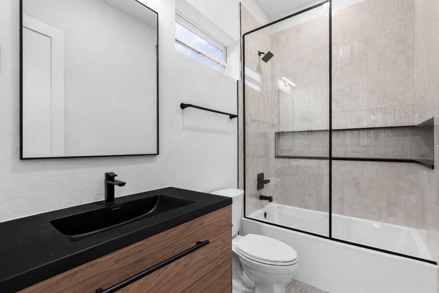 Crisp and composed, thesecondary ensuite pairs a wood-veneer vanity with an integratedgraphite basin and satin-black fittings. Vertically stacked tile and an elongated inset niche lend architectural order to the tub/shower, while a clerestory window introduces soft daylight—delivering a guest-ready bath that feels thoughtfully tailored.