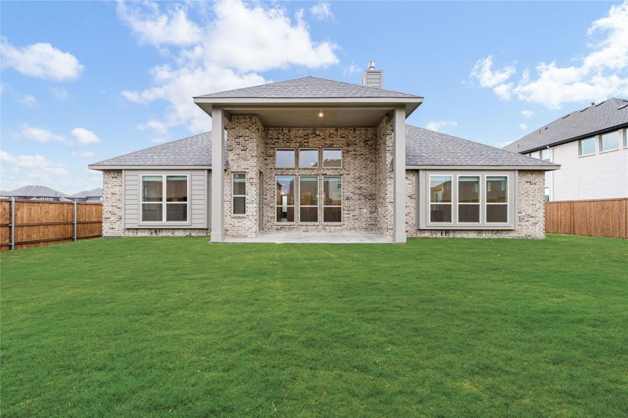 Rear exterior of single-story brick home with covered patio, large windows, and fenced backyard with green lawn