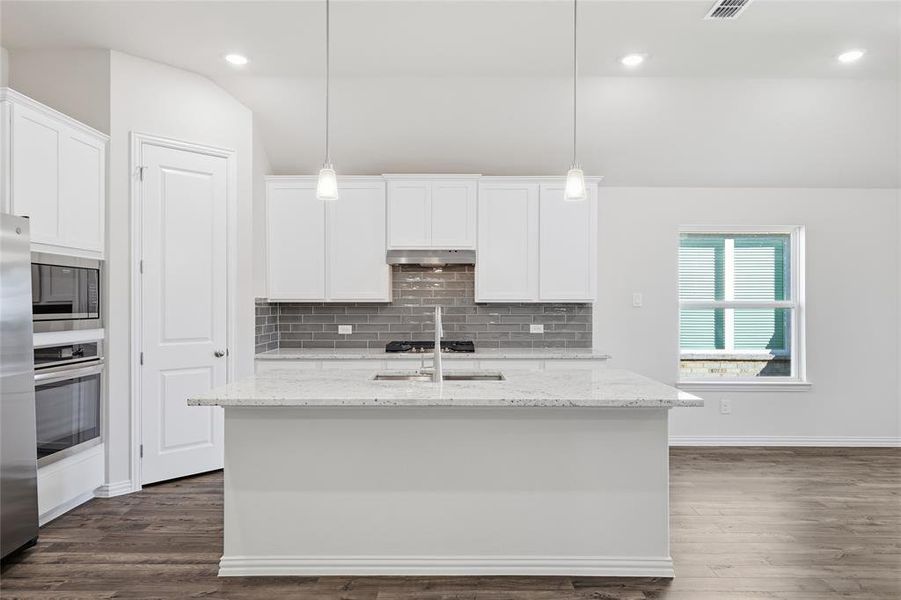 Spacious kitchen featuring an expansive central island with a sink, white cabinetry, integrated stainless steel appliances, a grey subway tile backsplash, and wood-finish flooring