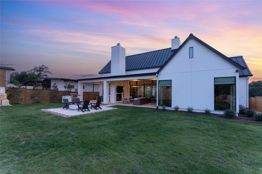 Spacious backyard featuring a manicured lawn, an outdoor fire pit area with stone surround, a covered patio with recessed lighting, and a modern white exterior with a dark metal roof Spacious backyard featuring a manicured lawn, an outdoor fire pit area with stone surround, a covered patio with recessed lighting, and a modern white exterior with a dark metal roof