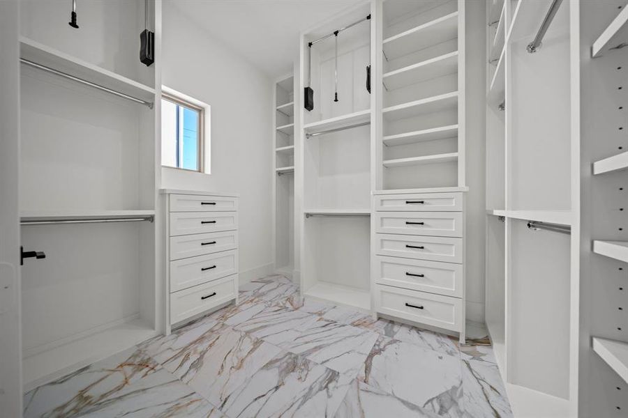 This spacious primary walk-in closet is a dream come true, offering custom-built shelving, ample drawer space, and pull-down rods for added convenience and organization. The elegant design features marble-style flooring and abundant natural light, creating a luxurious and functional storage solution. Perfectly tailored for all your wardrobe needs, this closet is both stylish and practical.