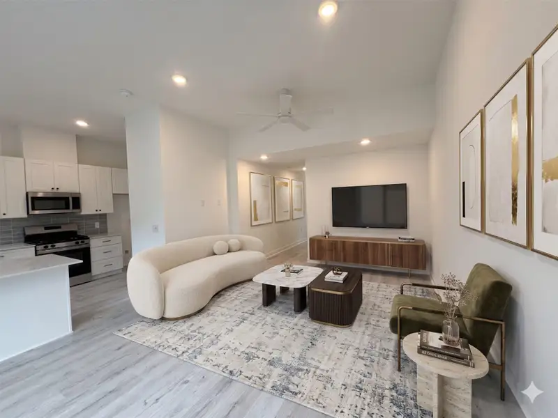 This modern living area features an open layout with a sleek kitchen, light wood flooring, and contemporary furnishings. The space is bright with recessed lighting and a ceiling fan, creating a welcoming and stylish environment.