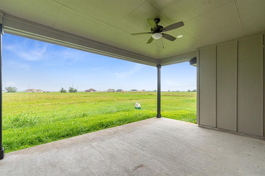 Oversized covered patio with a ceiling fan, overlooking expansive green fields, offering a serene outdoor living space with a view.